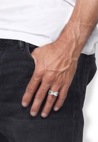 Sølvflettet ring båret på en hånd med synlige årer, sat mod en ensfarvet hvid baggrund, med sort denimjeans delvist synlige.