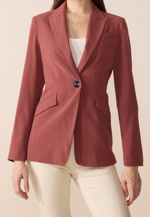 Mujer con blazer ajustada de color óxido con un solo botón, bolsillos con solapa y pantalones beige, de pie frente a un fondo liso.