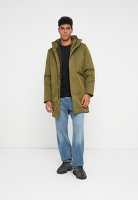 Olivgrüne Parka mit einer strukturierten, mit Fell gefütterten Kapuze und großen Seitentaschen, getragen über einem schwarzen Hemd und abgenutzten blauen Jeans.