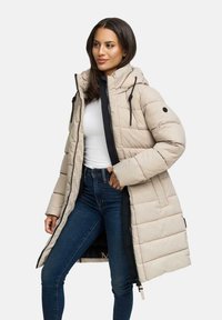 Vrouw in een beige lange gewatteerde jas met capuchon, witte shirt en blauwe spijkerbroek, staande met hand in de zak, kijkend opzij.