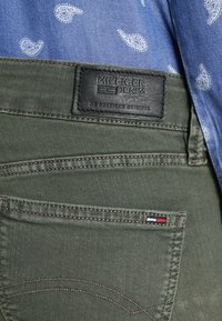 Tejido de denim verde oliva, parche con "HILFIGER DENIM," costuras marrones en contraste, pequeño acento en rojo, blanco y azul cerca del bolsillo. Camisa azul con patrón.