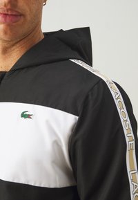 Man som bär en svart och vit Lacoste hoodie med en grön krokodil-logotyp och varumärkestape på axeln och ärmen.