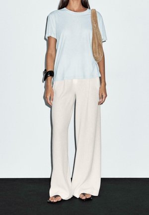 Kvinde iført lyseblå kortærmet skjorte, brede beige bukser, sort armbånd, tan skuldertaske og sorte sandaler, stående på mørkt gulv.