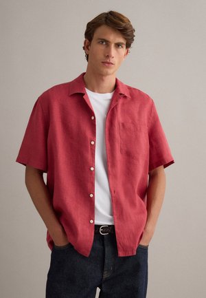 Junger Mann trägt ein rotes kurzärmeliges Hemd über einem weißen T-Shirt, mit dunklen Jeans und Händen in den Taschen, steht vor einer schlichten Wand.