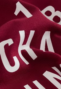 Punaruskea kangasta, jossa on suuria, rohkeita valkoisia kirjaimia. Pinnassa on sileä, jäykähkö kohokuvio, joka osoittaa pehmeän ja joustavan materiaalin.