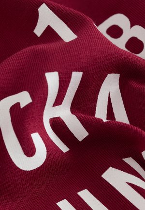 Punaruskea kangasta, jossa on suuria, rohkeita valkoisia kirjaimia. Pinnassa on sileä, jäykähkö kohokuvio, joka osoittaa pehmeän ja joustavan materiaalin.