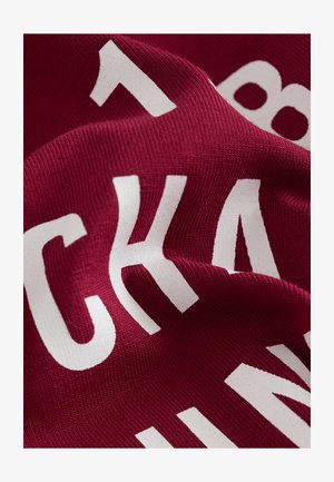 Punaruskea kangasta, jossa on suuria, rohkeita valkoisia kirjaimia. Pinnassa on sileä, jäykähkö kohokuvio, joka osoittaa pehmeän ja joustavan materiaalin.