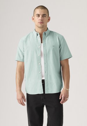 Homme portant une chemise boutonnée à manches courtes vert clair par-dessus un débardeur blanc et un pantalon noir, debout devant un fond clair uni.