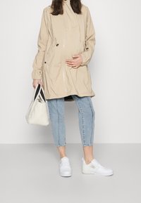 Beige voděodolná bunda s kapucí, elastickými rukávy, dvěma kapsami, v kombinaci se světle modrými džíny a bílými teniskami, a bílou taškou.