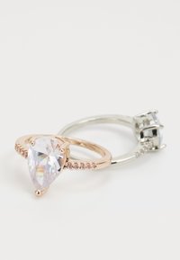 Dos anillos: uno presenta una piedra preciosa clara en forma de pera engastada en oro rosa con piedras más pequeñas a lo largo de la banda; el otro es de plata con una piedra preciosa en forma de cuadrado.