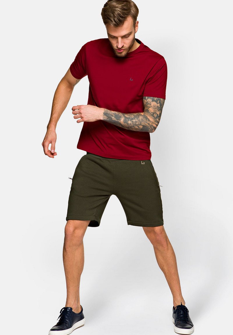 Czerwona koszulka z krótkim rękawem i okrągłym dekoltem z małym logo. Połączona z oliwkowymi szortami z zamykanymi kieszeniami. Nosi się z granatowymi trampkami.