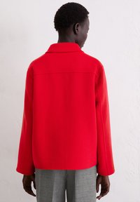 Rode wollen jas met kraag, voorzien van lange mouwen, een losse pasvorm en een rechte zoom. Gedragen over grijze broek, gezien vanaf de achterkant.