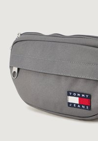 Pochette in tessuto grigio con chiusura a zip, dotata di tasca frontale e una patch con il logo Tommy Jeans in blu, rosso e bianco.