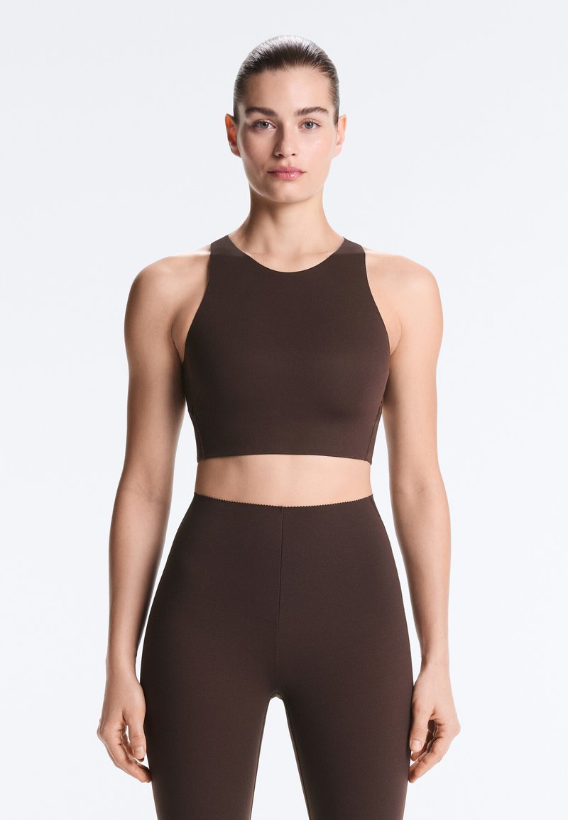 Femme aux cheveux lissés en arrière portant un crop top marron foncé sans manches et un legging taille haute assorti, debout devant un fond blanc uni.