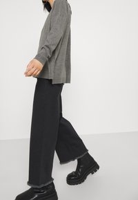 Grauer Strickpullover kombiniert mit weit geschnittenen schwarzen Jeans mit fransigen Säumen und klobigen schwarzen Stiefeln, die einen lockeren Sitz und einen Kontrast in der Textur hervorheben.