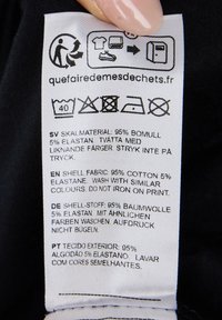Clothing care label with multilingual instructions, white fabric with black text. Details include washing icons and material composition: 95% cotton, 5% elastane.