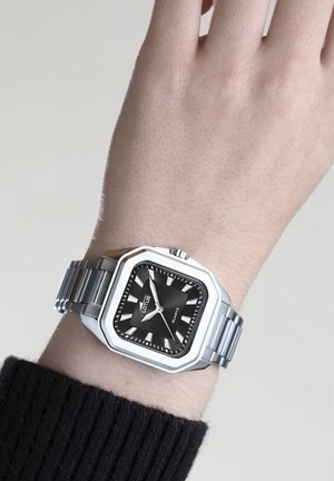 Silberfarbene Edelstahluhr mit schwarzem quadratischem Ziffernblatt, weißen Stundenmarkierungen und einem Metallarmband. Marke „Lotus“ auf dem Ziffernblatt angezeigt.