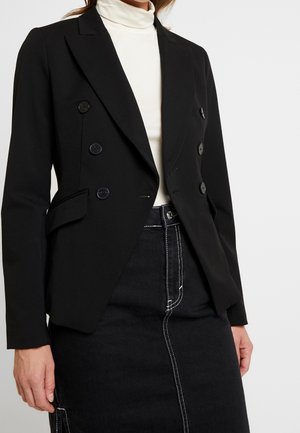 Donna che indossa un blazer doppiopetto nero sopra un dolcevita bianco e una gonna nera con dettagli di cuciture bianche.