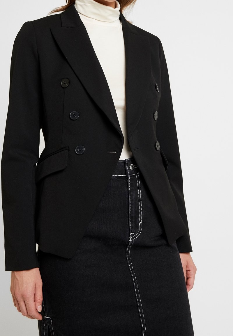 Femme portant un blazer noir croisé sur un col roulé blanc et une jupe noire avec des détails de couture blancs.