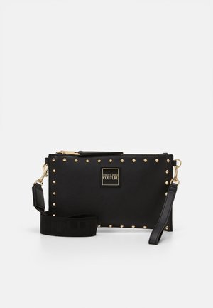 Pochette rectangulaire noire avec des clous dorés sur les bords, étiquette Versace Jeans Couture et sangle noire détachable avec des éléments métalliques dorés.