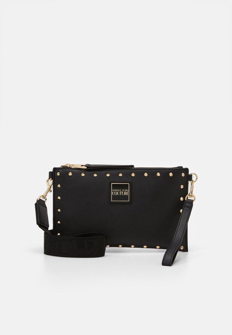 Pochette rectangulaire noire avec des clous dorés sur les bords, étiquette Versace Jeans Couture et sangle noire détachable avec des éléments métalliques dorés.