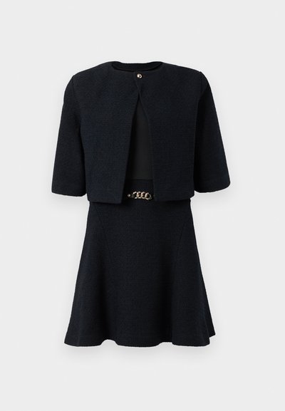 Černé šatky z twillu s krátkými rukávy, včetně zkrácené bundy a rozšířené sukně, doplněné zlatým páskem v pase.