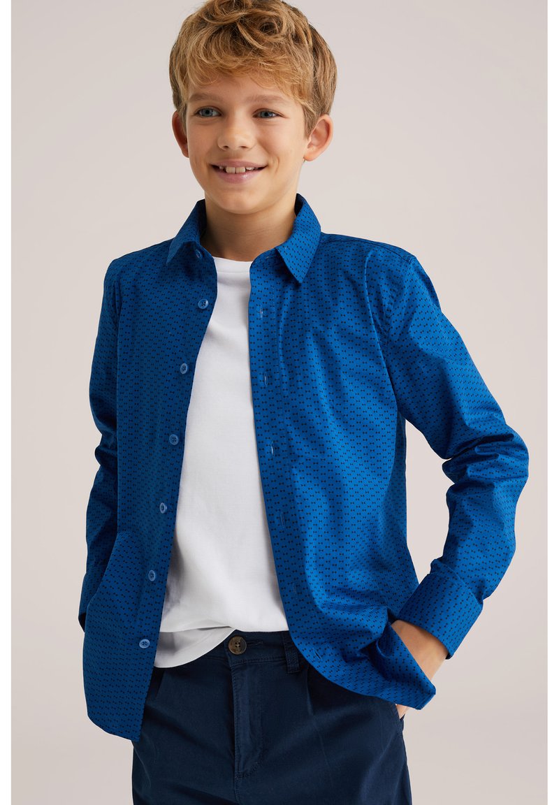 Blå skjorta med knappar och ett mönster av små svarta prickar, långa ärmar och krage, bärs över en vit t-shirt, ihop med mörka byxor.