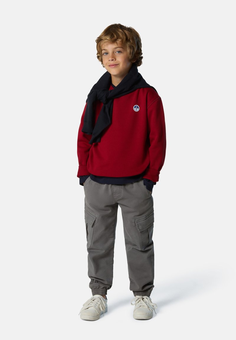 Felpa rossa con dettagli blu navy, pantaloni cargo grigi con polsini elasticizzati e scarpe da ginnastica bianche. Una sciarpa blu navy è appoggiata sulle spalle.