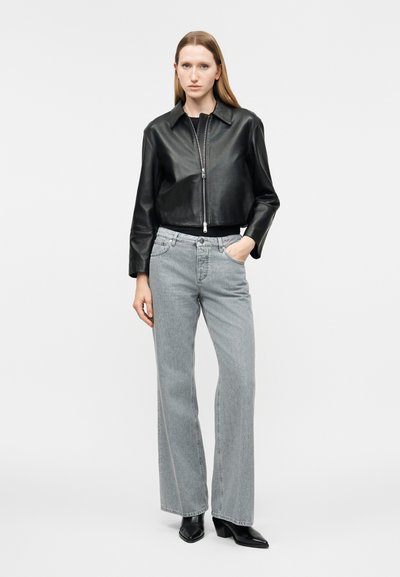 Γυναίκα που φοράει μαύρο κοντό δερμάτινο μπουφάν, ανοιχτό γκρι ψηλόμεσα φαρδιά τζιν και μαύρες μυτερές μπότες, στέκεται με το ένα χέρι στην τσέπη.