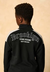 Chłopiec z krótkimi kręconymi włosami, ubrany w czarną i szaro-skalną kurtkę z napisem "Brooklyn Future Systems Twenty Four Seven" z tyłu.