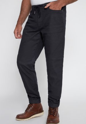 Pantalones de chándal negros con cinturilla elástica y cordón, que cuentan con bolsillos laterales y puños ajustados, combinados con botas marrones con cordones.
