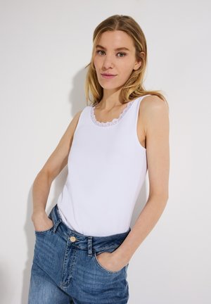 Woman with blonde hair wearing white sleeveless top with lace trim and blue jeans, standing with hands in pockets against white background.