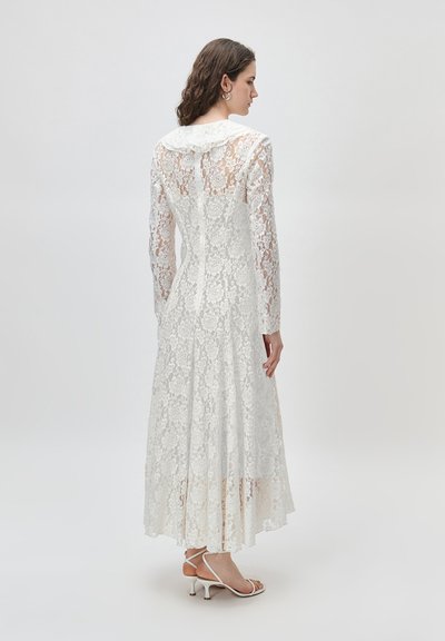 Γυναίκα που φοράει μακρύ λευκό δαντελένιο φόρεμα με μακριά μανίκια, γιακά και σχεδιασμό ανοιχτής πλάτης, συνδυασμένο με λευκά σανδάλια με τακούνι.