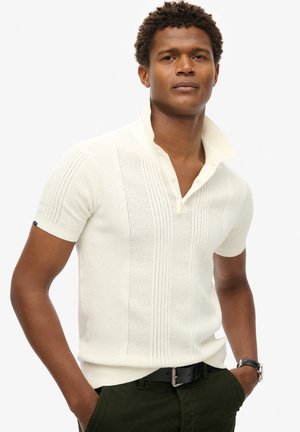 Short-sleeve, cream knit polo shirt with a high collar, featuring vertical stripes and a three-button placket. Worn with dark green pants.