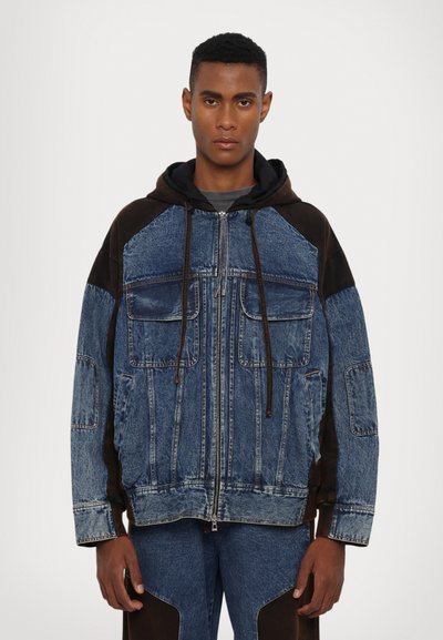 Denim jacket with brown fabric accents, front zipper, two large chest pockets, and a hood. Features patchwork design in varying shades of blue.