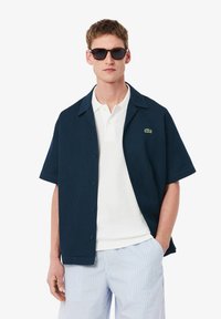 Μπλέ κοντομάνικο πουκάμισο με κουμπιά και υφή, φορεμένο πάνω από λευκό πόλο, σε συνδυασμό με ανοιχτόχρωμα ριγέ σορτς και γυαλιά ηλίου.