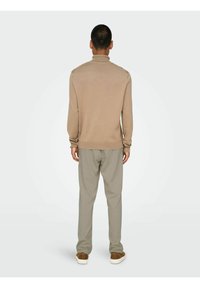 Beige strikpullover med høj krave, lange ærmer og ribbede manchetter, parret med løst siddende lysegrå bukser og brune sneakers.
