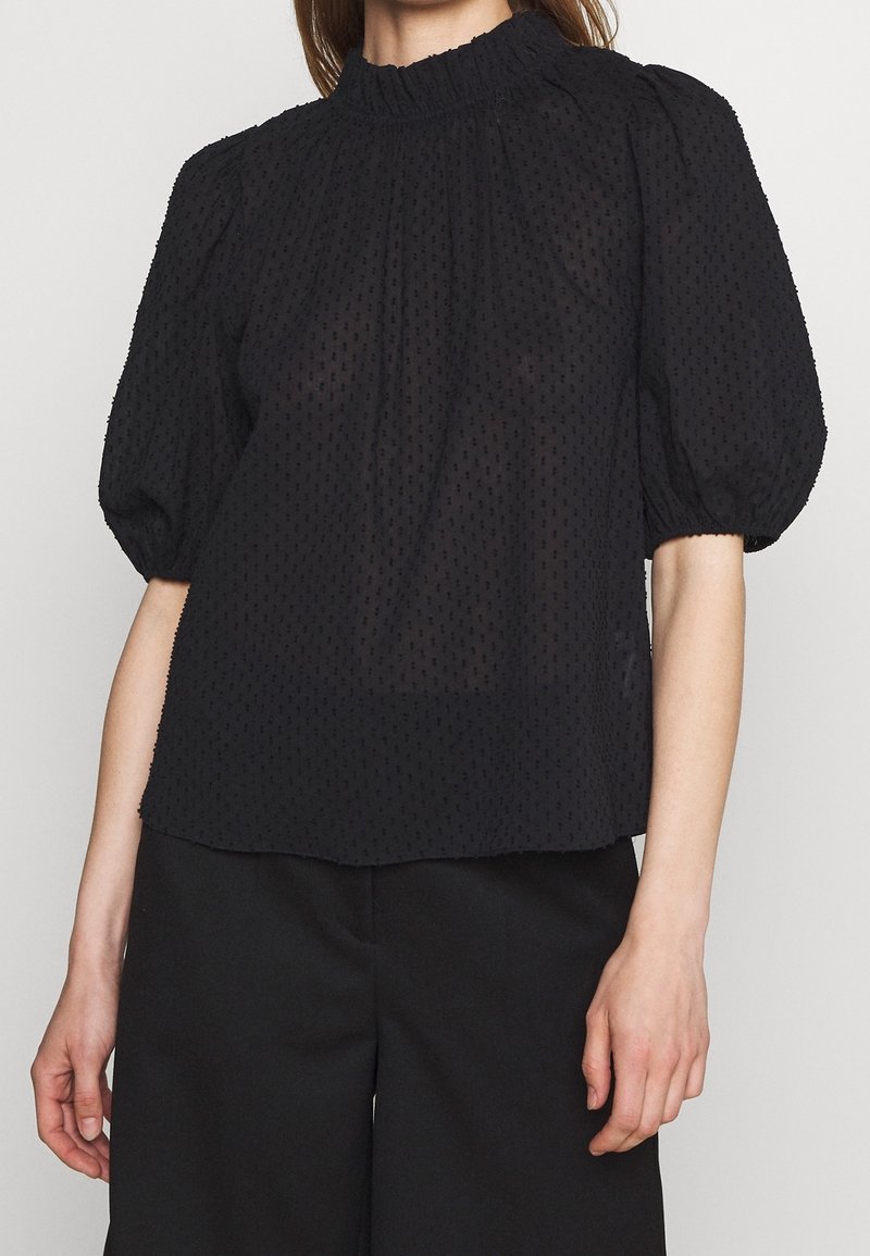 Femme portant un chemisier noir transparent texturé avec des manches courtes bouffantes et un col montant à volants, associé à un pantalon noir.