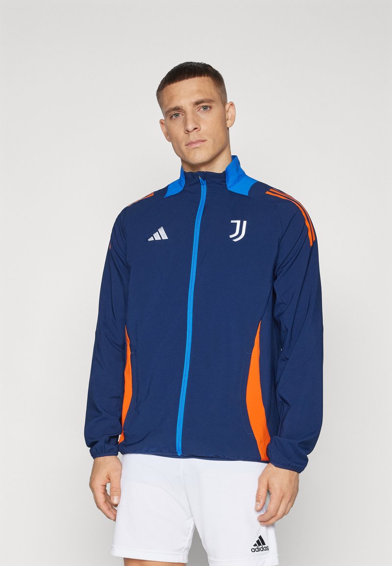 Giacca della Marina con dettagli arancioni e blu, chiusura frontale a zip, colletto alto e tasche laterali. Presenta i loghi di adidas e Juventus. Materiale leggero.