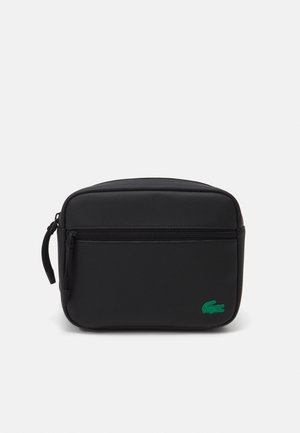 Pochette noire compacte zippée avec une poche avant, une poignée latérale et un logo crocodile vert dans le coin inférieur droit.