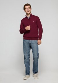 Suéter burdeos con media cremallera, puños y dobladillo acanalados, combinado con jeans azul claro y zapatillas color beige. El modelo sonríe, con las manos en los bolsillos.