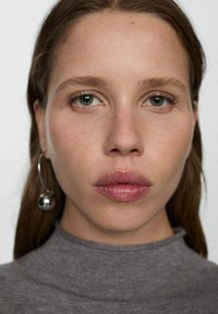 Gros plan d'un visage avec des cheveux châtains mi-longs, une peau claire et des yeux verts marquants. Porte un col roulé gris et une grande boucle d'oreille circulaire.