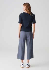 Marineblauw kortmouwtop met een gegolfde zoom, gecombineerd met grijze wijde broek en witte sneakers met blauwe accenten. Model gezien vanuit de achterkant.