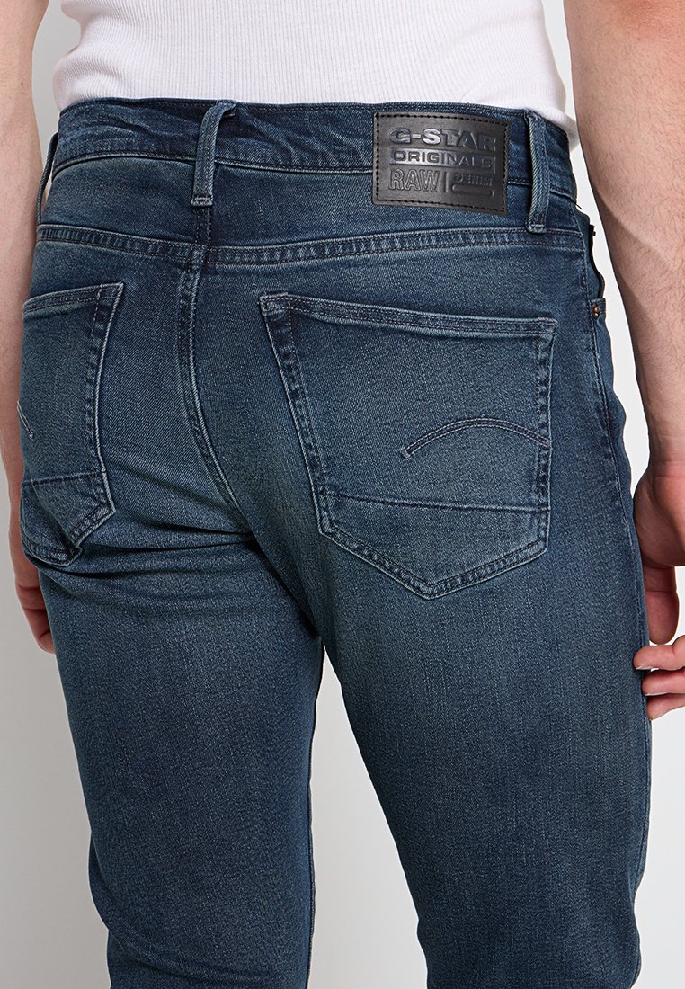 Jean en denim bleu avec une coupe slim, des poches arrière avec un design subtil, une étiquette en cuir à la taille et une finition délavée.