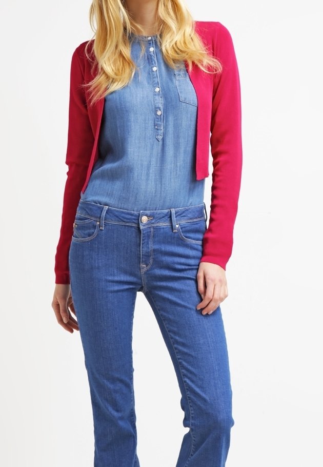 Femme portant une chemise chambray bleue boutonnée, un jean en denim bleu et un cardigan court rose vif, posant avec une main près de la hanche.