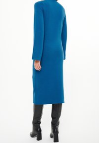 Långärmad, teal klänning i texturerad tyg med rak silhuett och hög halsringning, kombinerad med knähöga svarta stövlar.