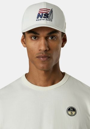 Uomo che indossa un cappellino da baseball bianco con il logo "NS North Sails" e una maglietta bianca con girocollo con un piccolo emblema circolare "North Sails" sul petto.