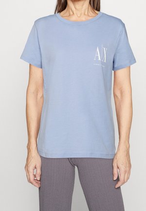 Persoon draagt een lichtblauw Armani Exchange T-shirt met korte mouwen en een wit logo op de linkerborst, gecombineerd met geribbelde grijze broek.