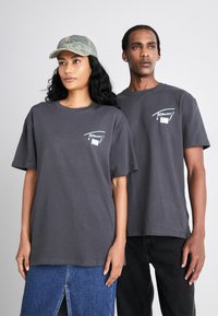 Camisetas de algodón gris usadas por dos personas, con un pequeño gráfico en la parte frontal y mangas cortas. Una persona lleva una gorra de camuflaje.
