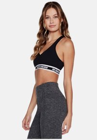 Sujetador deportivo negro con una banda de marca blanca, que presenta un diseño de escote en V y tirantes anchos, combinado con leggins grises de textura alta.